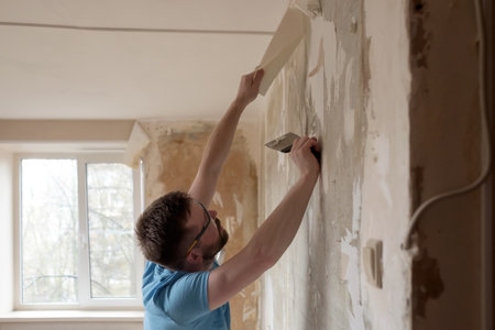 Hardworking man removes old wallpaper from the wall with a spatula in the room. Home repairs.の写真素材