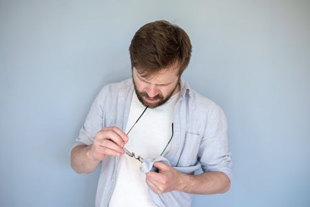 Concentrated Caucasian man cleans glasses from dust and dirt, wiping them with his shirt. Gray background.の写真素材