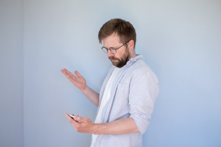 Puzzled bearded man in glasses and casual clothes looks at smartphone, he is confused and makes a hand gesture. Gray background.の写真素材