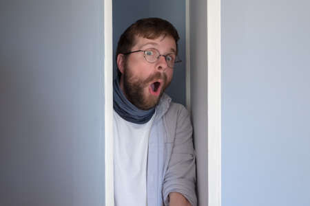 Bearded man in casual clothes is hiding in a closet, he peeps through the open door and looks in surprise with an open mouth and big eyes.の写真素材