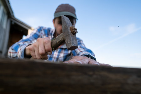 Carpenter hands make a bench by hammering nails into a plank in a village yard against a blue sky.の写真素材