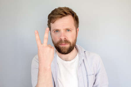 Serious bearded man in a shirt shows with fingers the number two or a symbol of victory. Gray background.の写真素材