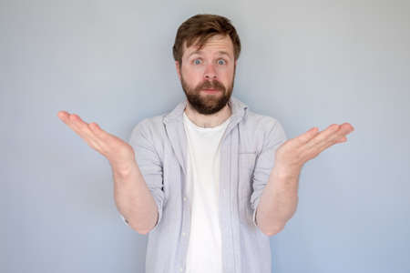 Bewildered bearded man in a shirt, shocked, throws up hands to the sides and stares dumbfounded. Gray background.の写真素材