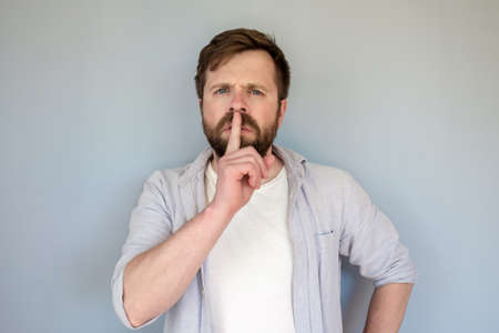 Serious man put his index finger to lips and demands silence, looking sternly at the camera. Gray background.の写真素材