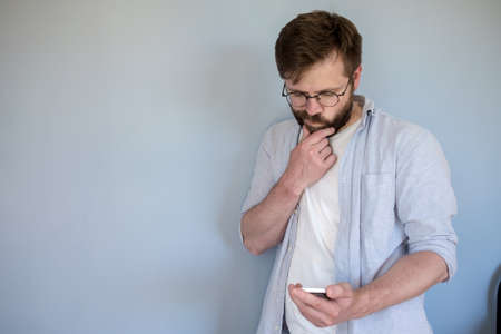 Pensive man with glasses looks at a smartphone, he is confused and holds chin with hand. Copy space. Gray background.の写真素材