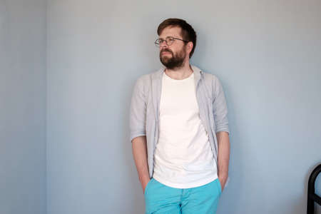 Attractive bearded man in glasses and casual clothes looks pensively to the side, standing against a gray wall in a room.の写真素材
