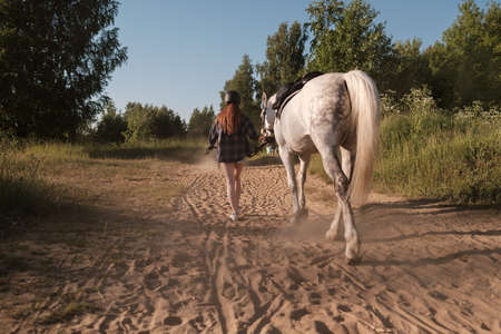 Woman in a helmet walks with her horse along a dusty sandy road, holding her by the bridle, at sunset, against the background of the sky.の写真素材