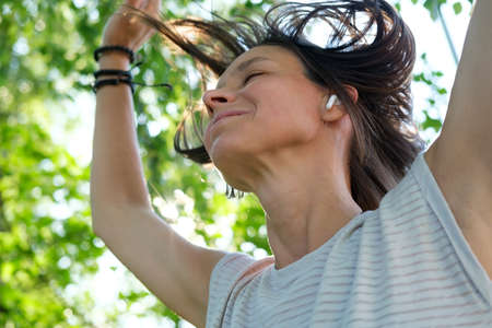 Happy woman listens to music in wireless headphones and dances, with her hair flying up, on a summer sunny day.の写真素材