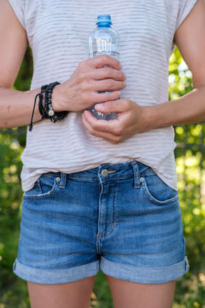 Woman holds a bottle of cool drinking water in her hands, she presses it to her body, on a summer day, against a blurred background.の写真素材