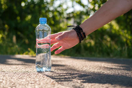 Female hand takes the bottle with cool drinking water, standing on the asphalt road on a sunny summer day.の写真素材