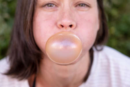 Closeup portrait of funny women, carefully inflates bubble gum, with narrowed eyes and round cheeks, outdoors.の写真素材