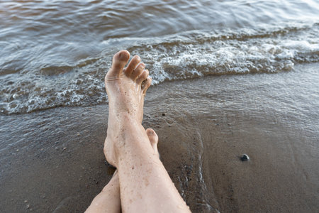 Wet female feet on a sandy beach near the water. Woman is resting while sitting by the lake, on a summer day. Lifestyle.の写真素材