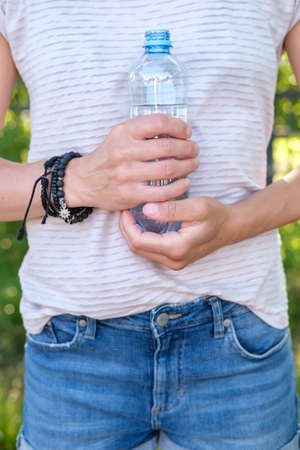 Woman holds a bottle of cool drinking water in her hands, she presses it to her body, on a summer day. Close-up.の写真素材