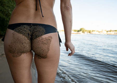 Woman ass in the sand, in a black swimsuit, on the banks of the river, on a city beach, on a blurred background.の写真素材