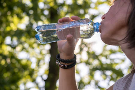 Woman drinks cool drinking water from a plastic bottle on a hot summer day against the backdrop of trees and the sky. Close-up.の写真素材