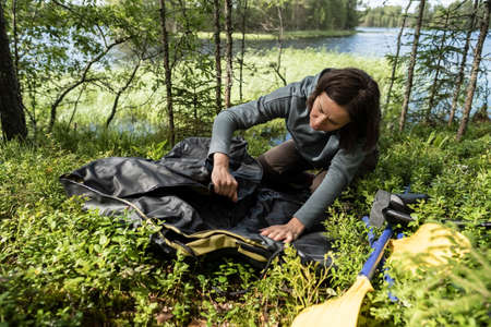 Woman unfolds a new rubber boat, oars are lying nearby, against the backdrop of a lake. Preparing for a boat trip or fishing.の写真素材