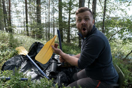 Shocked man holds a broken oar he found while unpacking a new rubber boat against the backdrop of a lake. Preparing for a boat trip.の写真素材