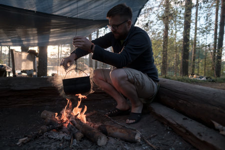 Tourist cooking dinner over a campfire at the camp. Man stirring food in a bowler, which hangs over a fire, on a summer evening.の写真素材