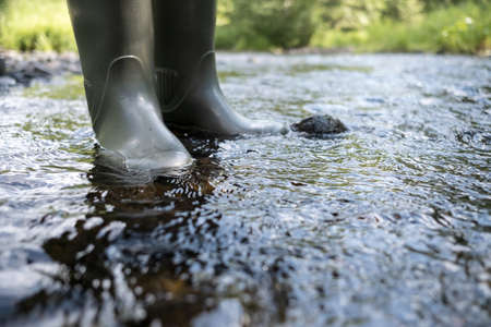 Male feet in waterproof rubber boots walk along the river, against the backdrop of greenery, on a summer day.の写真素材