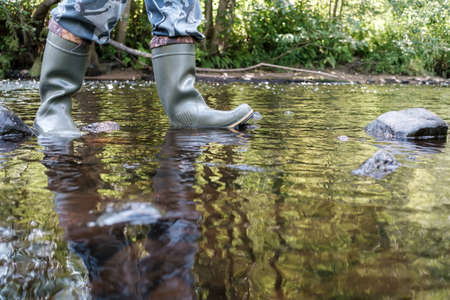 Male feet in waterproof rubber boots walk along the river, against the backdrop of greenery, on a summer day.の写真素材