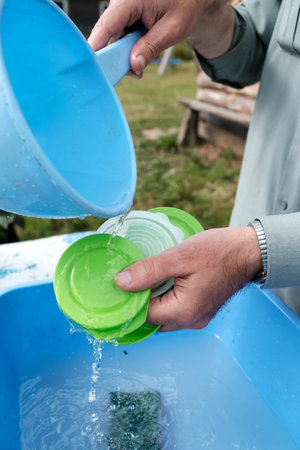 Male hands pour clean water from a ladle onto plastic lids to rinse off the detergent in a country yard, outdoors. Rural traditions.の写真素材