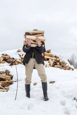 Man is carrying an armload of chopped wood in a rustic courtyard, on a frosty winter day. Heating season in the village.の写真素材
