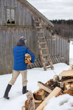 Man carries an armful of firewood, against the backdrop of a barn, in the countryside, on a winter day. Heating season in the village.の写真素材