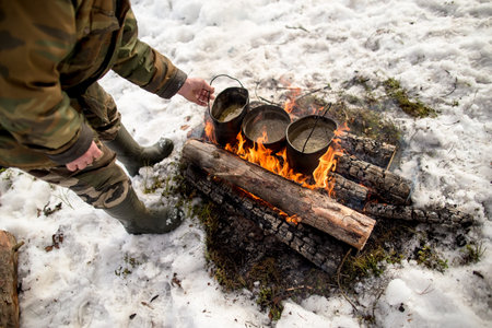 Three pots with boiling water on a campfire, a man in warm clothes is standing next to him, on a winter day. Cooking on the hike.の写真素材