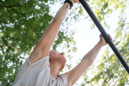 Woman hangs on a horizontal bar, on the background of sky and trees. Healthy and sporty lifestyle. Bottom view.の写真素材