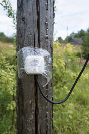 Electrical socket with a wire, protected from moisture by a transparent visor, is attached to an old wooden post, outdoors.の写真素材