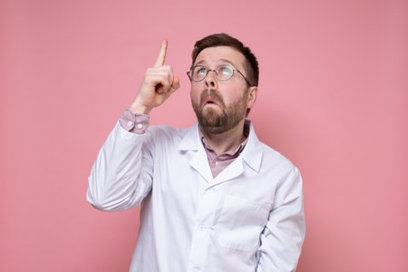 Surprised doctor in a white coat and glasses points his finger up and looks puzzled. Pink background.の写真素材