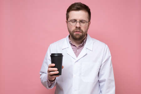 Doctor in a white coat and glasses is very tired and looks sleepy, he holds a paper cup of coffee in hands and stands with eyes closed.の写真素材