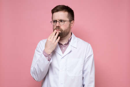Caucasian doctor in a white coat pensively and anxiously looks to the side, holding hand to face. Pink background.の写真素材