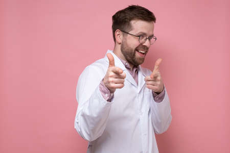 Funny doctor in a white coat is joking and smiling pointing at someone with fingers. Pink background.の写真素材