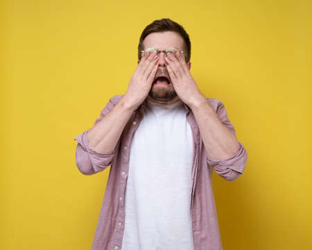 Unhappy Caucasian man is upset and crying, he wipes eyes with hands and opens mouth in sobs. Yellow background.の写真素材