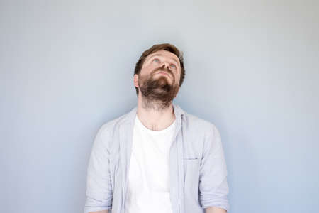 Unhappy, tired Caucasian bearded man in a shirt raised head and looked up, hands down in hopelessness. Gray background.の写真素材