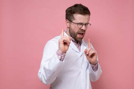 Disgruntled doctor in a white coat scolds someone, pointing strictly with fingers. Pink background.の写真素材