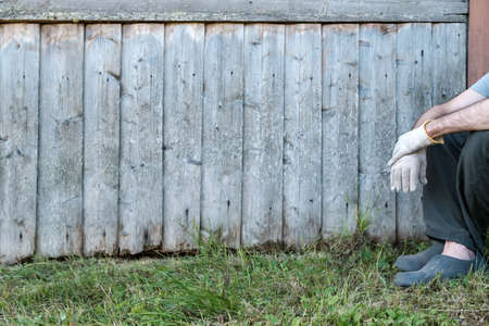 Old wooden wall of a house near which a man in work gloves is resting, in a village courtyard. Copy...