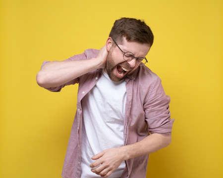 Man is experiencing severe pain in the neck area, he holds hand to the inflamed area and screams with eyes closed.の写真素材