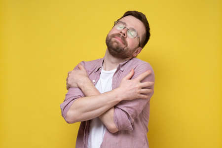Cute, bearded man with glasses gently hugs himself and dreamily closes eyes. Yellow background.の写真素材
