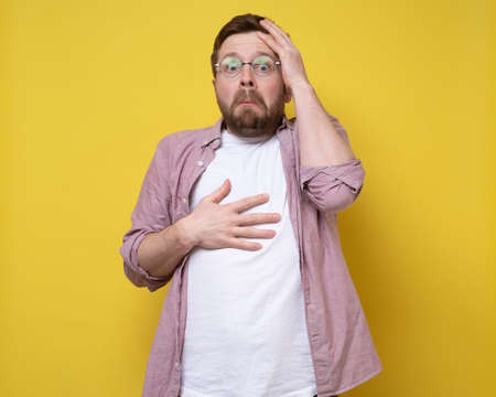 Shocked man with a frightened expression on face holds head and chest with hand in surprise. Yellow background.の写真素材