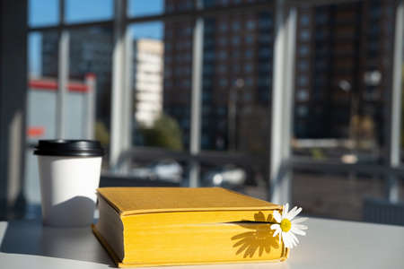 Closed yellow book with a bookmark with a chamomile flower and aromatic coffee in a disposable paper cup lie on the table in a cafe.の写真素材