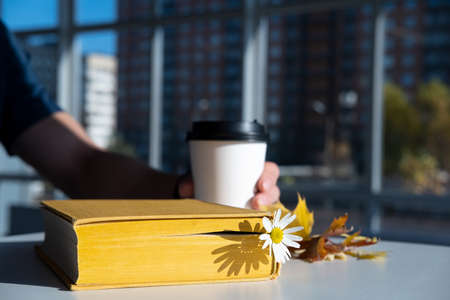 Book with a bookmark with a chamomile flower lies on the table, against the background a male hand holds a disposable cup of coffee.の写真素材