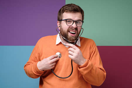Strange bearded man listens to his heart with a stethoscope, opening his mouth and looking at the camera.の写真素材