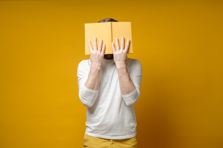 Student in a white t-shirt reads enthusiastically, covering his face with a book. Yellow background. Education concept.の写真素材