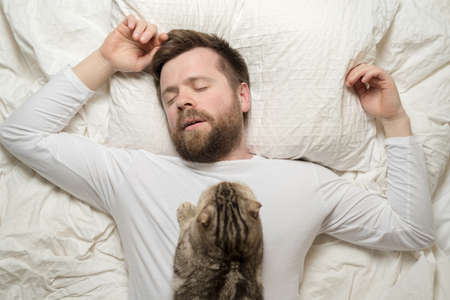 Man in white pajamas is asleep, and his adorable cat lies on his chest. Top view.の写真素材