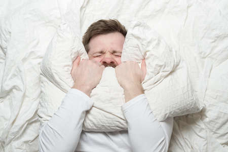Man in white pajamas is stressed, has insomnia, cannot sleep, nervously covers mouth with a pillow and squeezes eyes shut. Top view.の写真素材