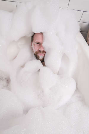 Satisfied man is almost completely covered with lush soap suds, he bathes in the bathroom, enjoys and relaxes. Top view.の写真素材