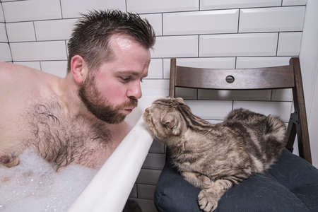 Man bathes in a bath in lush foam, next to him lies his adored cat, which is very interested in what is happening.の写真素材