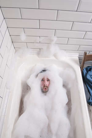 Man is bathing in a bathtub, he is covered in lush soap suds, only his face with a funny expression is visible.の写真素材